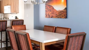 Un comedor con una mesa blanca y sillas tapizadas en marrón, una cocina con armarios de madera y un cuadro de un paisaje montañoso en la pared.