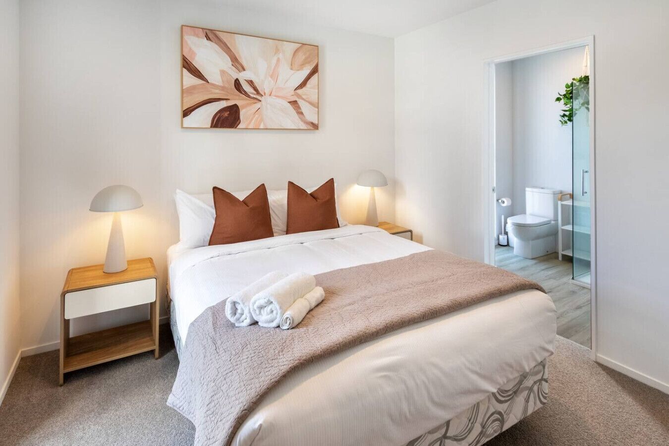 A hotel room with a bed, two bedside tables, a nightstand, a towel, a painting, and a bathroom visible through an open door.