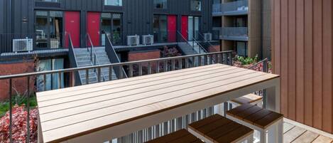 A modern outdoor seating area with a wooden table and benches, overlooking a building with balconies and red accents.