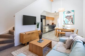 A modern apartment with a staircase, a living room with a sofa and coffee table, a dining area with a table and chairs, a kitchen with appliances, and a TV mounted on the wall. - Event Base Perfection + Balcony & Parking (Christchurch)