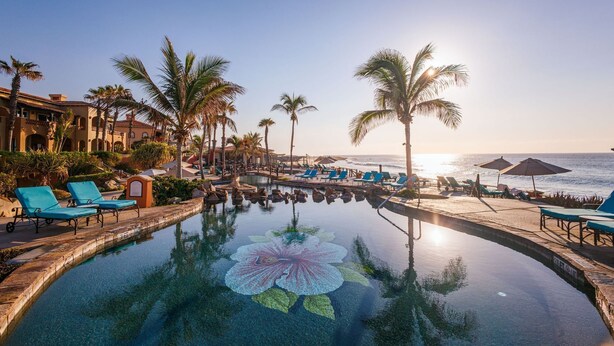 Une piscine de complexe hôtelier décorée de motifs de fleurs de lotus, entourée de palmiers et de chaises longues, et offrant une vue sur l’océan.