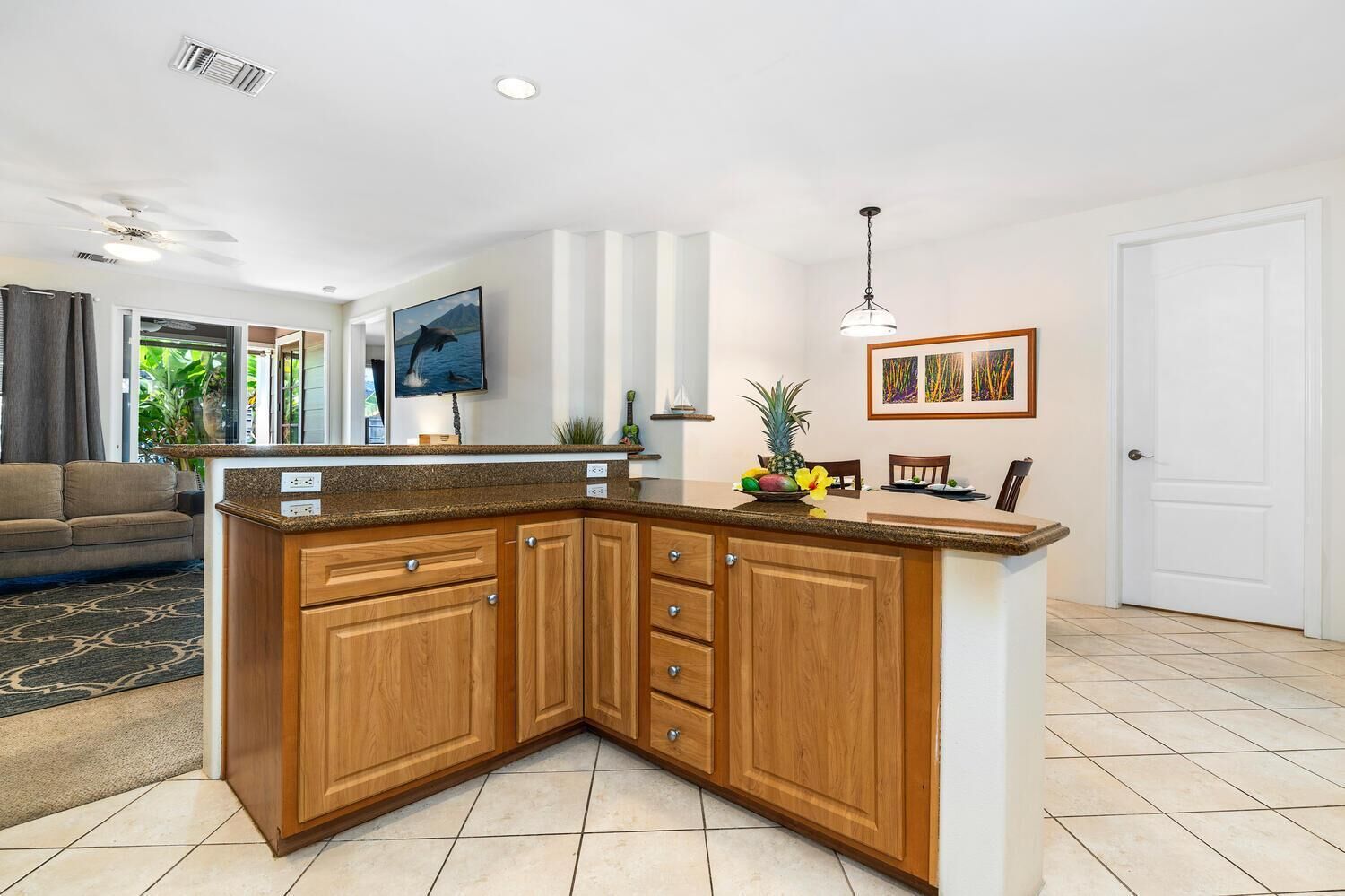 A kitchen with wooden cabinets, a granite countertop, a dining area with chairs, and a television on the wall.