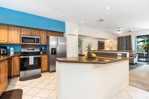 A modern kitchen with wooden cabinets, stainless steel appliances, a granite countertop island, and a view of the outdoor area. - Alii Cove EE3 Beautiful Ground Floor Unit With AC! (Kailua-Kona)