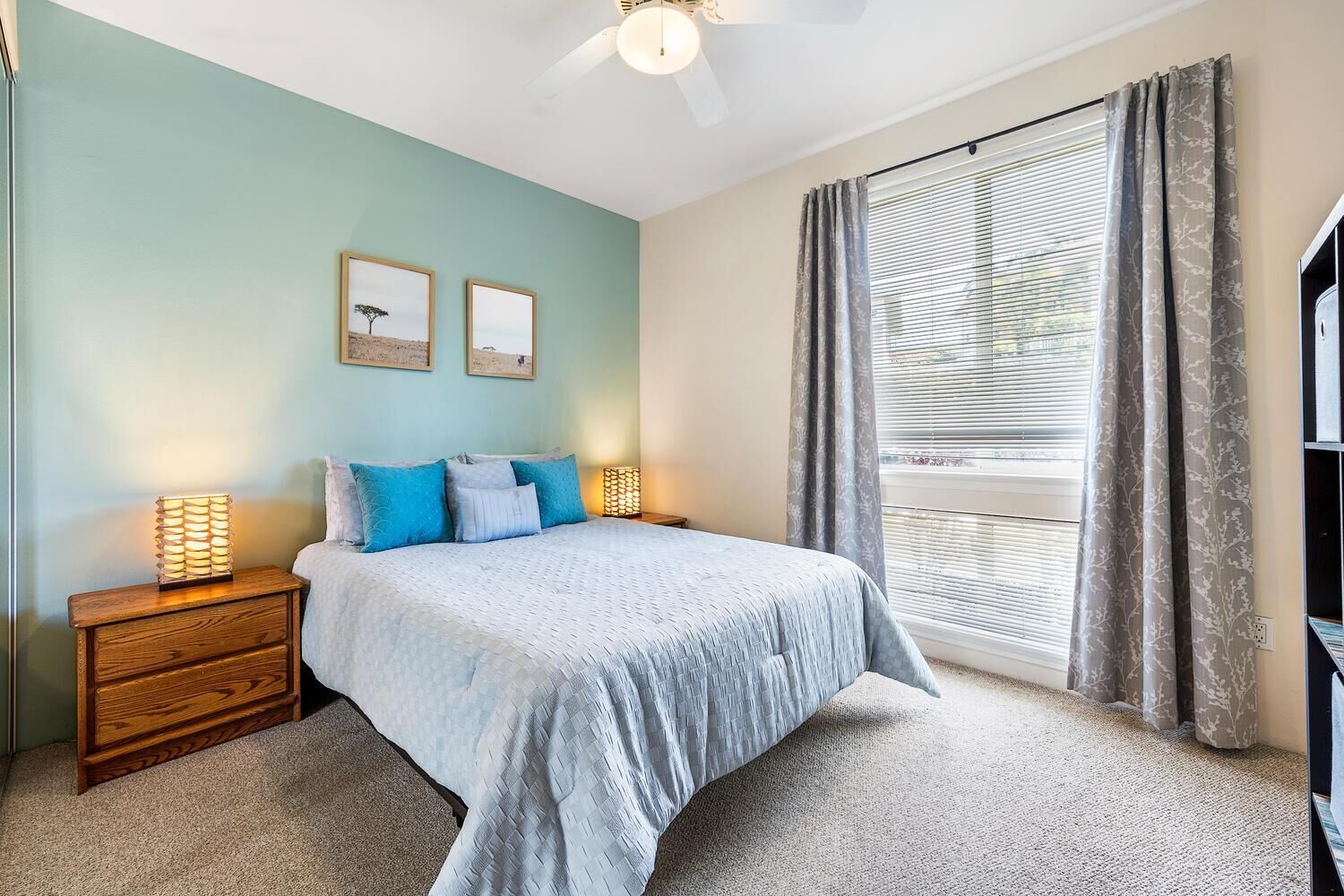 Une chambre à coucher comprenant un lit, deux tables de chevet, un ventilateur de plafond, une fenêtre avec des rideaux et des œuvres d’art murales.