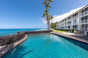 A swimming pool with a view of the ocean, a building with a balcony, and a stone wall. - Kona Magic Sands#305 (Kailua-Kona)