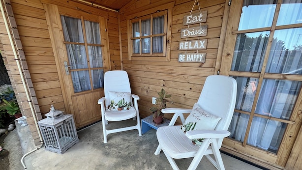 A wooden cabin with two white chairs, a small table, and a sign that reads 'Eat Drink Relax Be Happy.' - Cozy 1-bedroom cabin perfect for 2 in Willemstad (Willemstad)