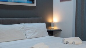 A neatly made bed with white linens and rolled towels, a gray headboard, and a wall-mounted lamp. - Deluxe Double Room 102 (Catania)