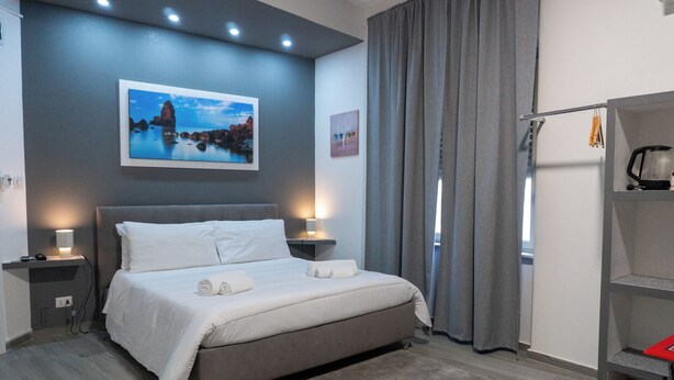 A modern bedroom with a bed, bedside lamps, a nightstand, a painting, a window with curtains, and a shelf with a kettle. - Deluxe Double Room 102 (Catania)