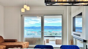 A modern living room with a sofa, dining table, chairs, and a large sliding glass door leading to a balcony with a view of a lake and buildings. - 2 bedroom awesome apartment in Turanj (Turanj)