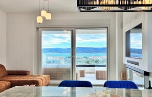 A modern living room with a sofa, dining table, chairs, and a large sliding glass door leading to a balcony with a view of a lake and buildings. - 2 bedroom awesome apartment in Turanj (Turanj)