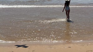Una playa con una persona volando una cometa, un velero a lo lejos e huellas en la arena.
