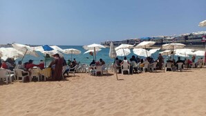 Una playa con hileras de sillas de plástico blancas y sombrillas, gente sentada y caminando, y una vista despejada del mar.