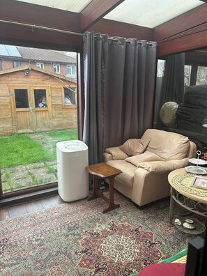 A cozy room with a leather sofa, a small wooden side table, a large window with curtains, and a patterned rug. - Cosy Family Home With Full Amenities (Uxbridge)