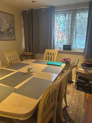 A dining area with a table set for six, surrounded by chairs, a radio, and a window with curtains. - Cosy Family Home With Full Amenities (Uxbridge)