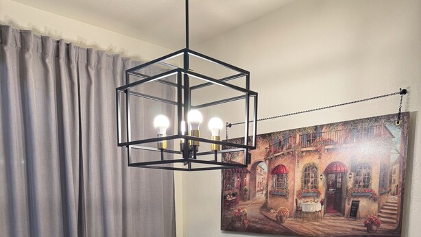 Un comedor con una mesa dispuesta para cuatro, sillas negras, una lámpara colgante moderna y un cuadro en la pared.