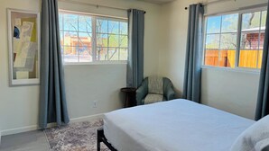 Un dormitorio con cama, dos ventanas con cortinas, una silla y una alfombra.