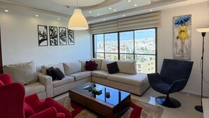 Ein modernes Wohnzimmer mit einer Eckcouch, einem roten Sessel, einem Glastisch und Blick auf die Stadt durch große Fenster.