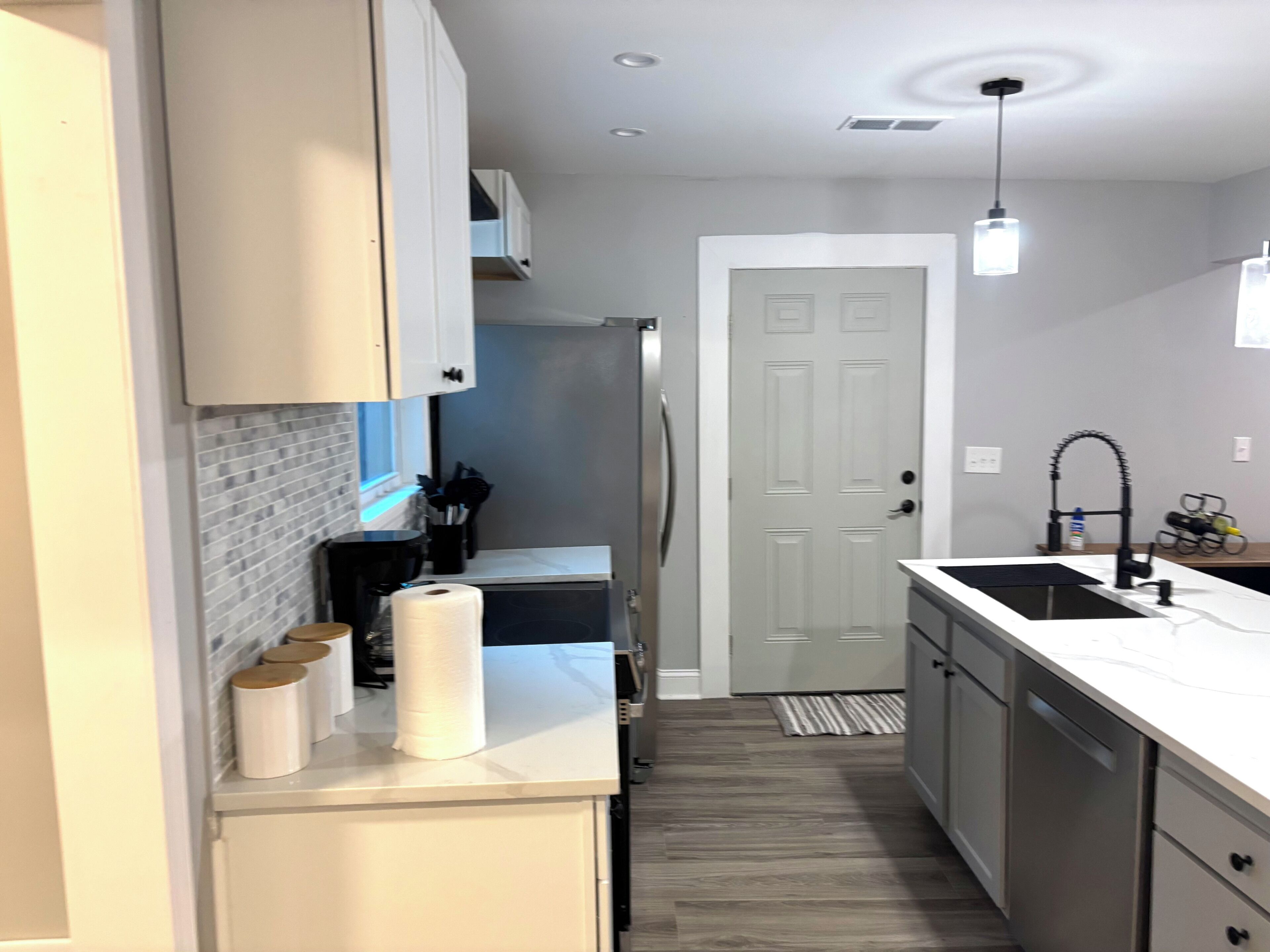 A modern kitchen with white cabinets, a stainless steel refrigerator, a built-in oven, a sink with a black faucet, and a wooden floor.