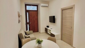 A modern living room with a round dining table, beige sofas, a small potted plant, a flat-screen TV, and a wooden door. - B&b SunHouse (Napoli)