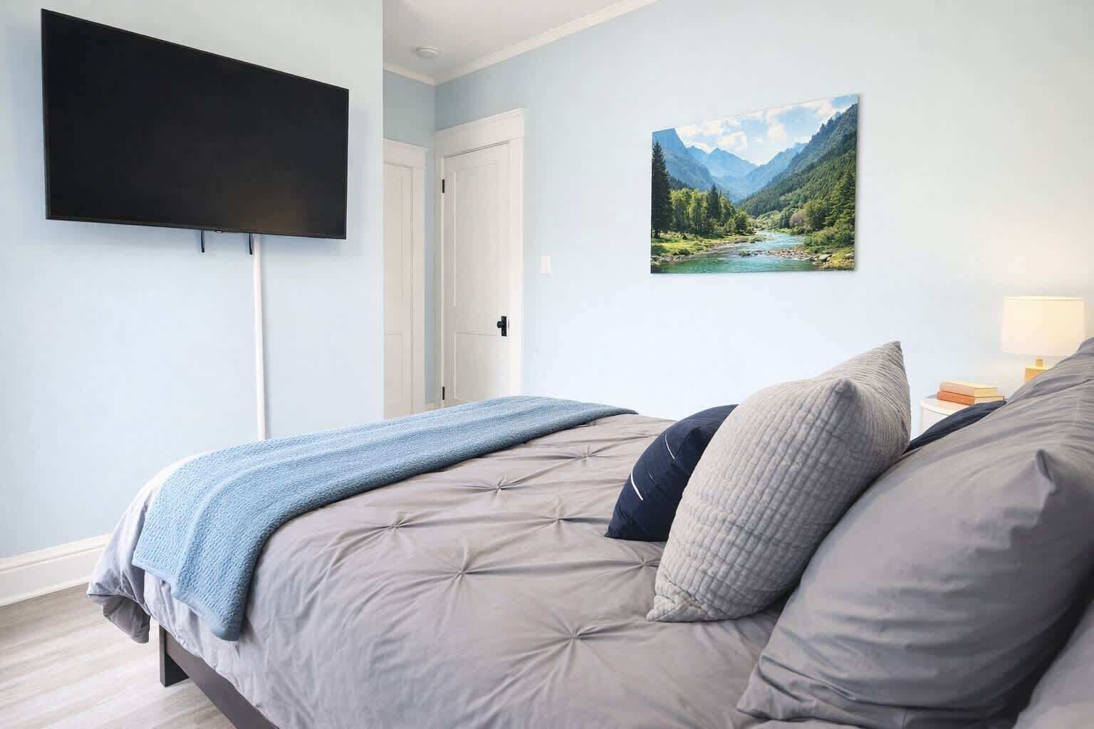 Un dormitorio con una cama, un televisor de pantalla plana montado en la pared, una puerta y un cuadro con un paisaje.