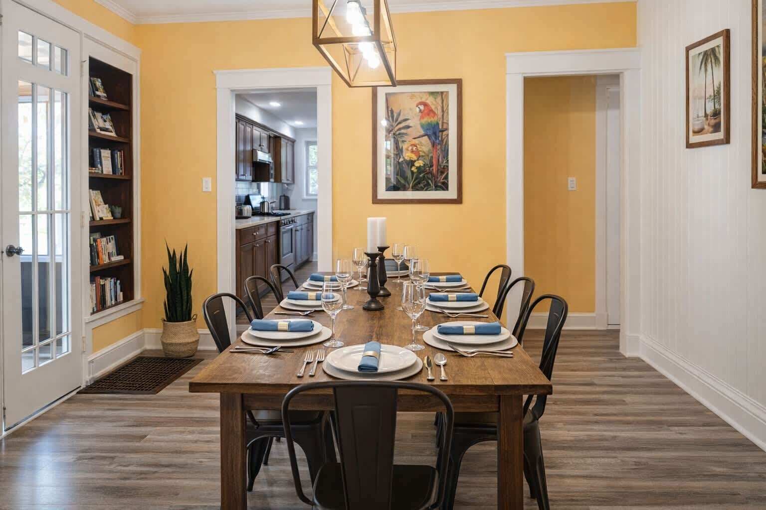 A dining room with a wooden table set for six, a kitchen in the background, and a bookshelf to the left.