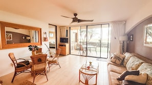 A spacious living room with a large sliding glass door leading to an outdoor area, a ceiling fan, and a television. - Lovely 2-bedroom condo with AC  Luana Kai A104 (Kihei)