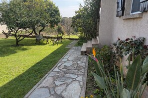 Un sendero de piedra lleva a una zona de estar con bancos, rodeada por un césped cuidado y árboles.