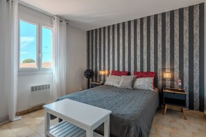 Ein Schlafzimmer mit einem Bett, einem Nachttisch, einem Standventilator und einem Fenster mit Vorhängen.