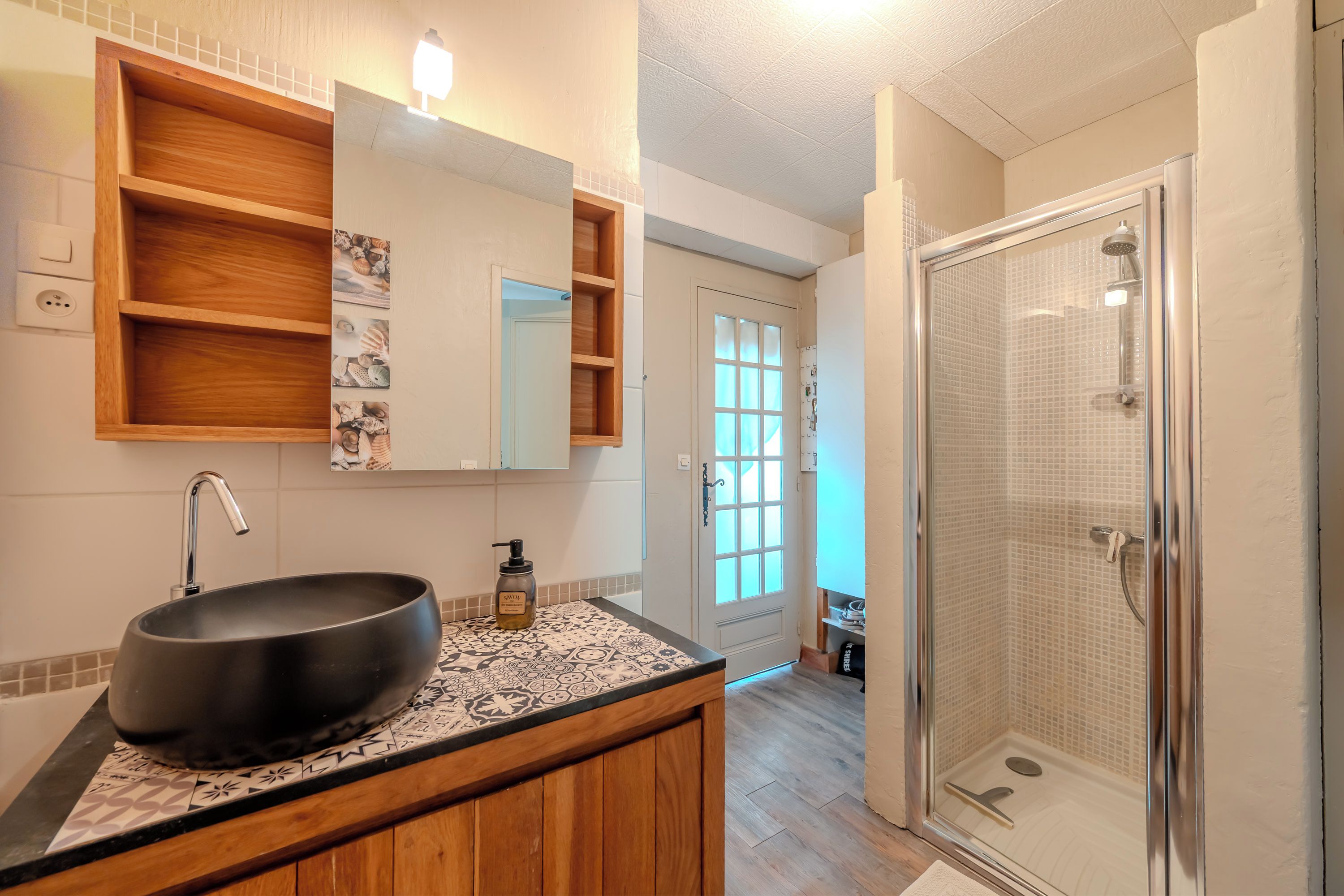Un baño con un lavabo negro grande, tocador de madera, un espejo con repisa arriba y zona de ducha con mampara de vidrio.