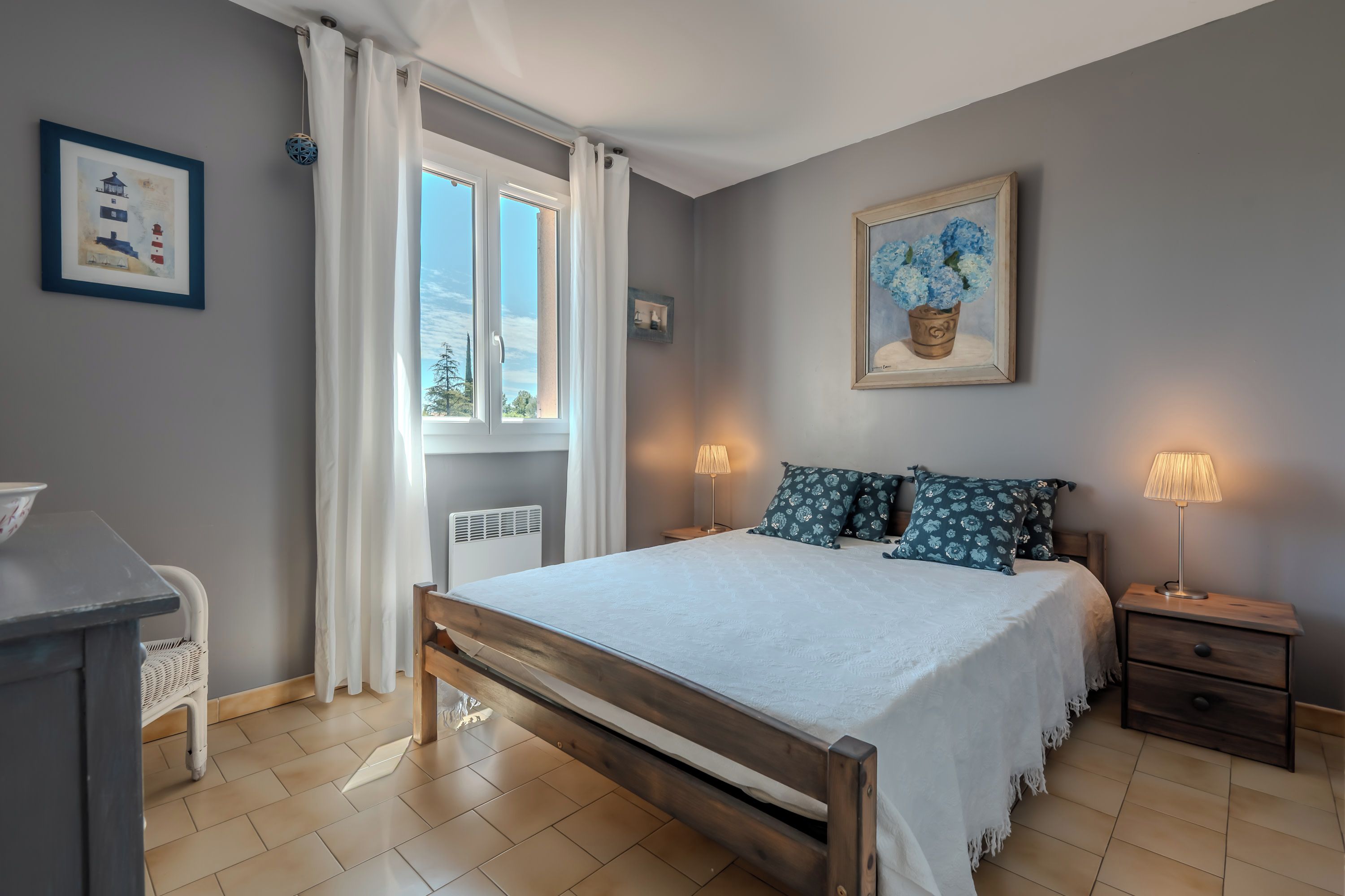 Une chambre à coucher avec un lit, des lampes de chevet, une table de nuit, une fenêtre avec des rideaux et un tableau encadré accroché au mur.