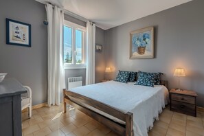 Een slaapkamer met een bed, nachtlampjes, een nachtkastje, een raam met gordijnen en een lijst met een schilderij aan de muur.