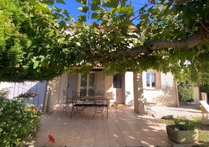 A patio with a table and chairs, surrounded by greenery and a building with windows. - La Maison Provençale - Piscine (L'Isle-sur-la-Sorgue)