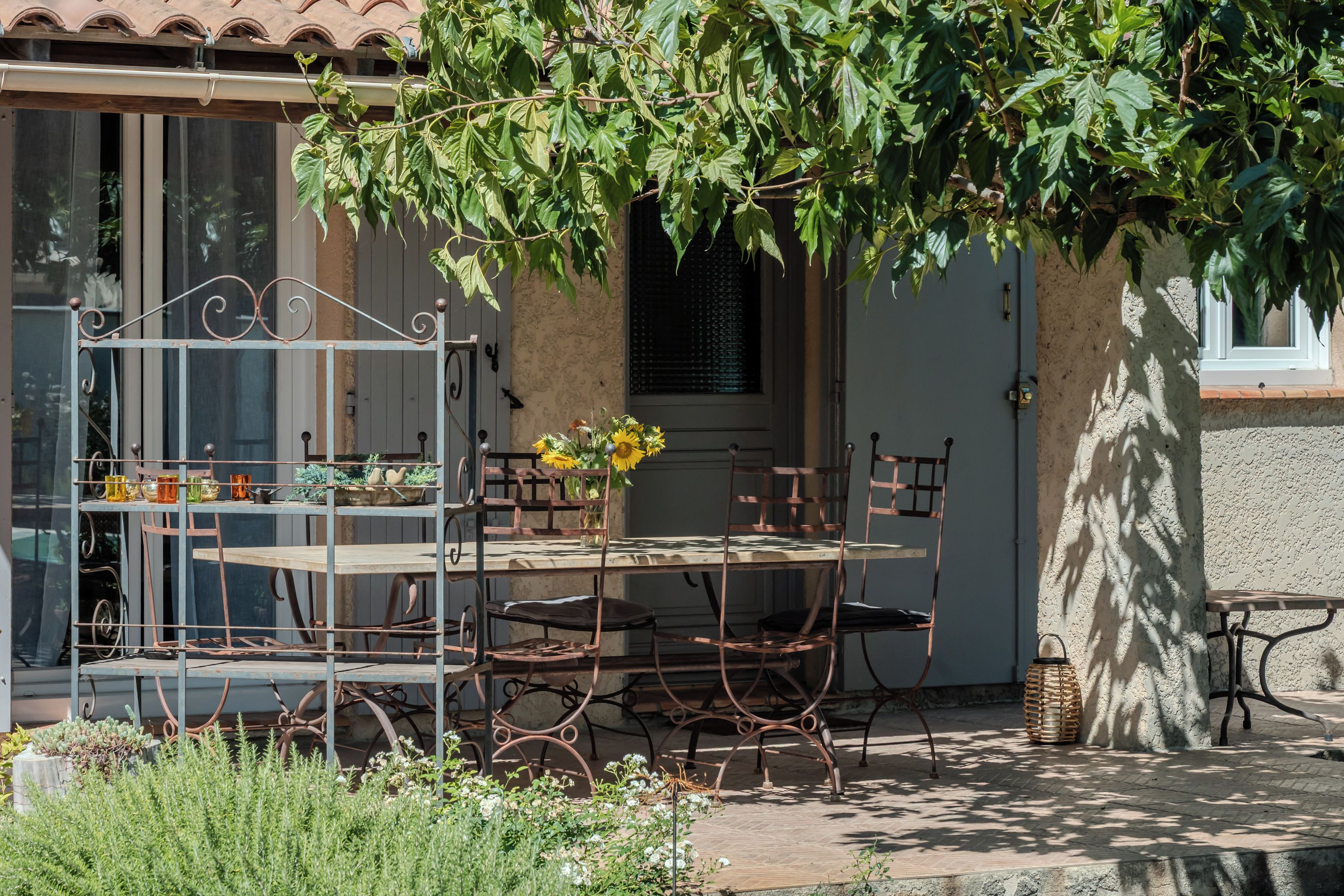 A rustic outdoor seating area with a metal table and chairs, surrounded by greenery and a building with a tiled roof.