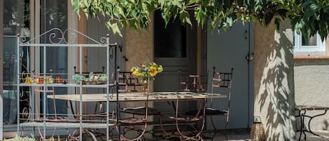 A rustic outdoor seating area with a metal table and chairs, surrounded by greenery and a building with a tiled roof.