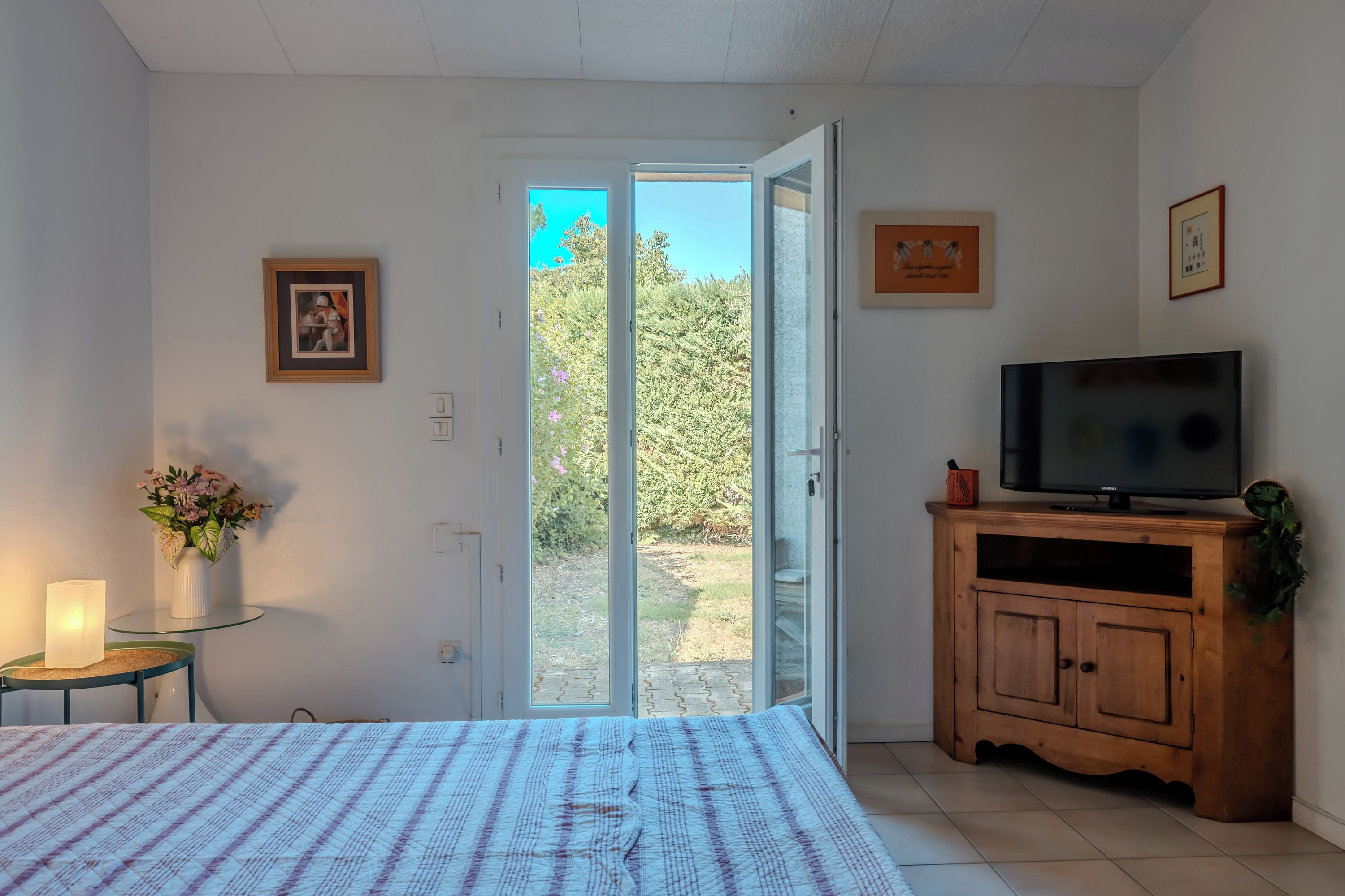 Un dormitorio con una cama, un televisor sobre un mueble de madera, un florero con flores y una puerta que da a un jardín.