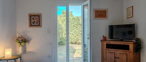 Quarto com uma cama, televisão em um armário de madeira, um vaso com flores e uma porta que dá para um jardim.