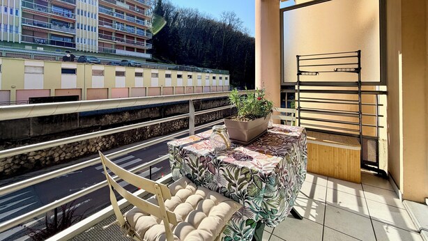 A balcony with a view of a train, a potted plant, a cushioned chair, and a small table. - T2 parking, prox thermes et centre-ville (Aix-les-Bains)