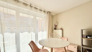 A dining area with a round table, two chairs, a radiator, a shelf with books, and a window with sheer curtains. - T2 parking, prox thermes et centre-ville (Aix-les-Bains)