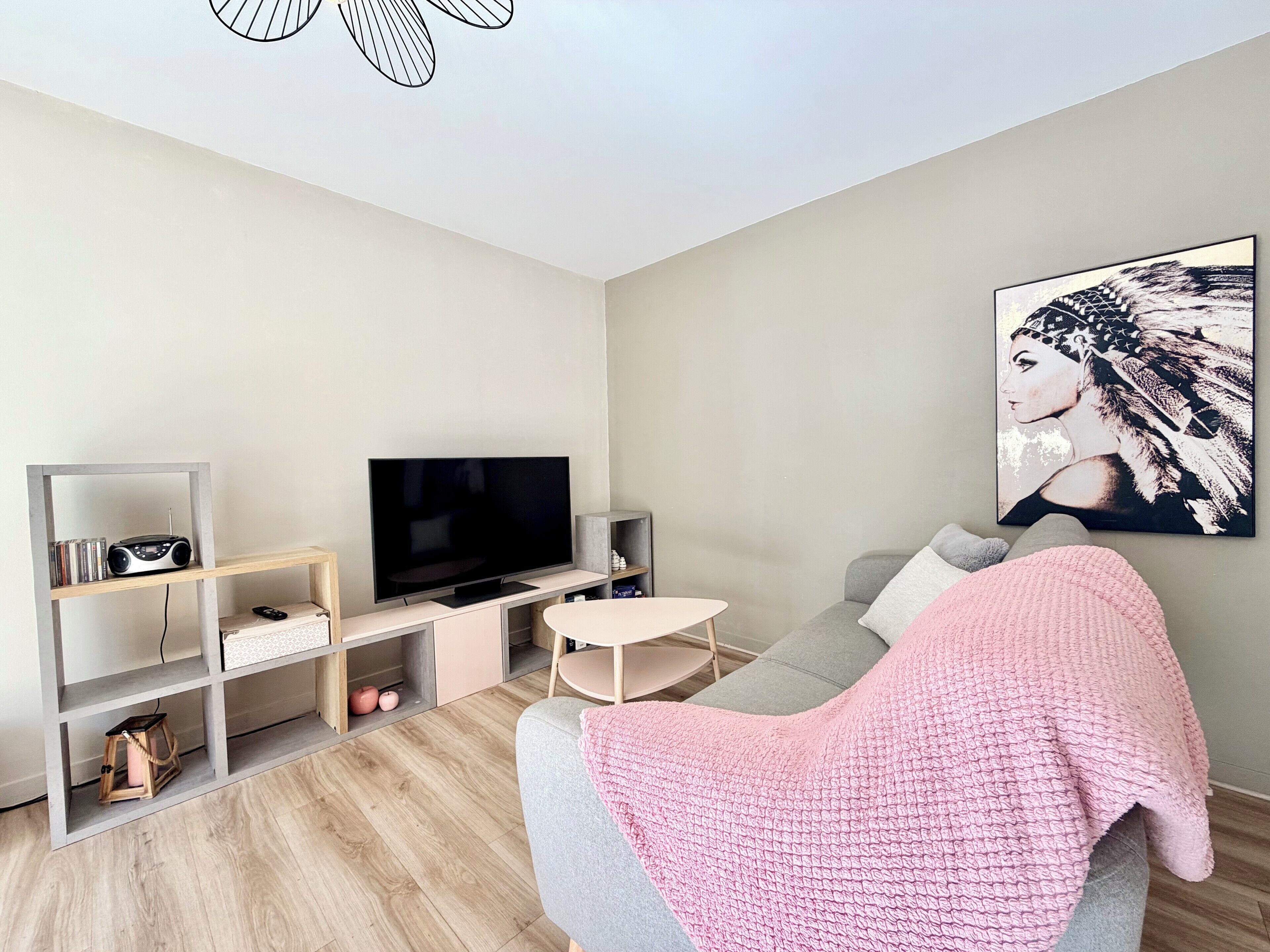 Ein modernes Wohnzimmer mit Flachbildfernseher, Holzböden, einem grauen Sofa, einer rosafarbenen Decke und einem an der Wand befestigten Kunstwerk.