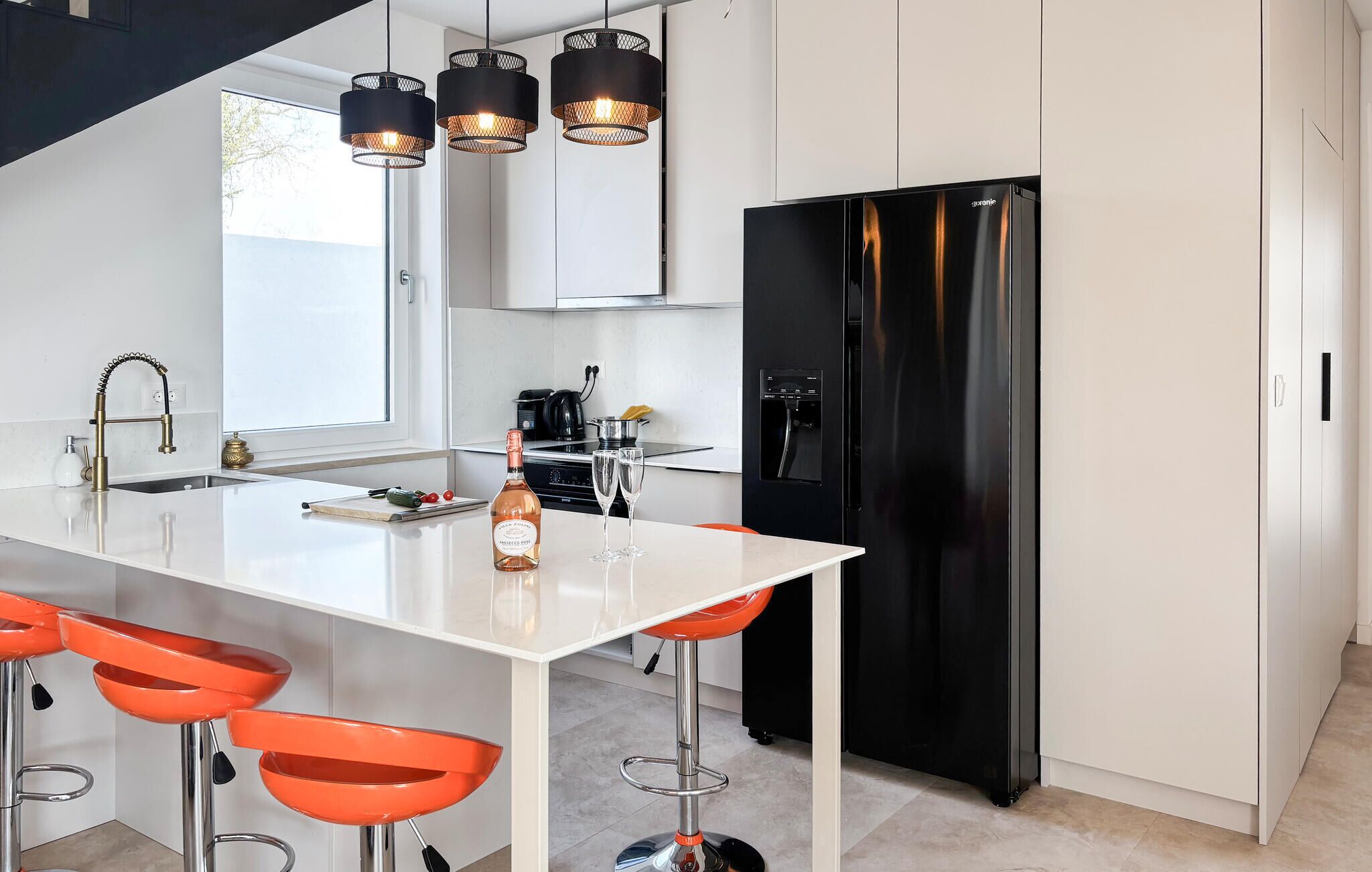 A modern kitchen with a white island, black refrigerator, orange bar stools, and pendant lights.