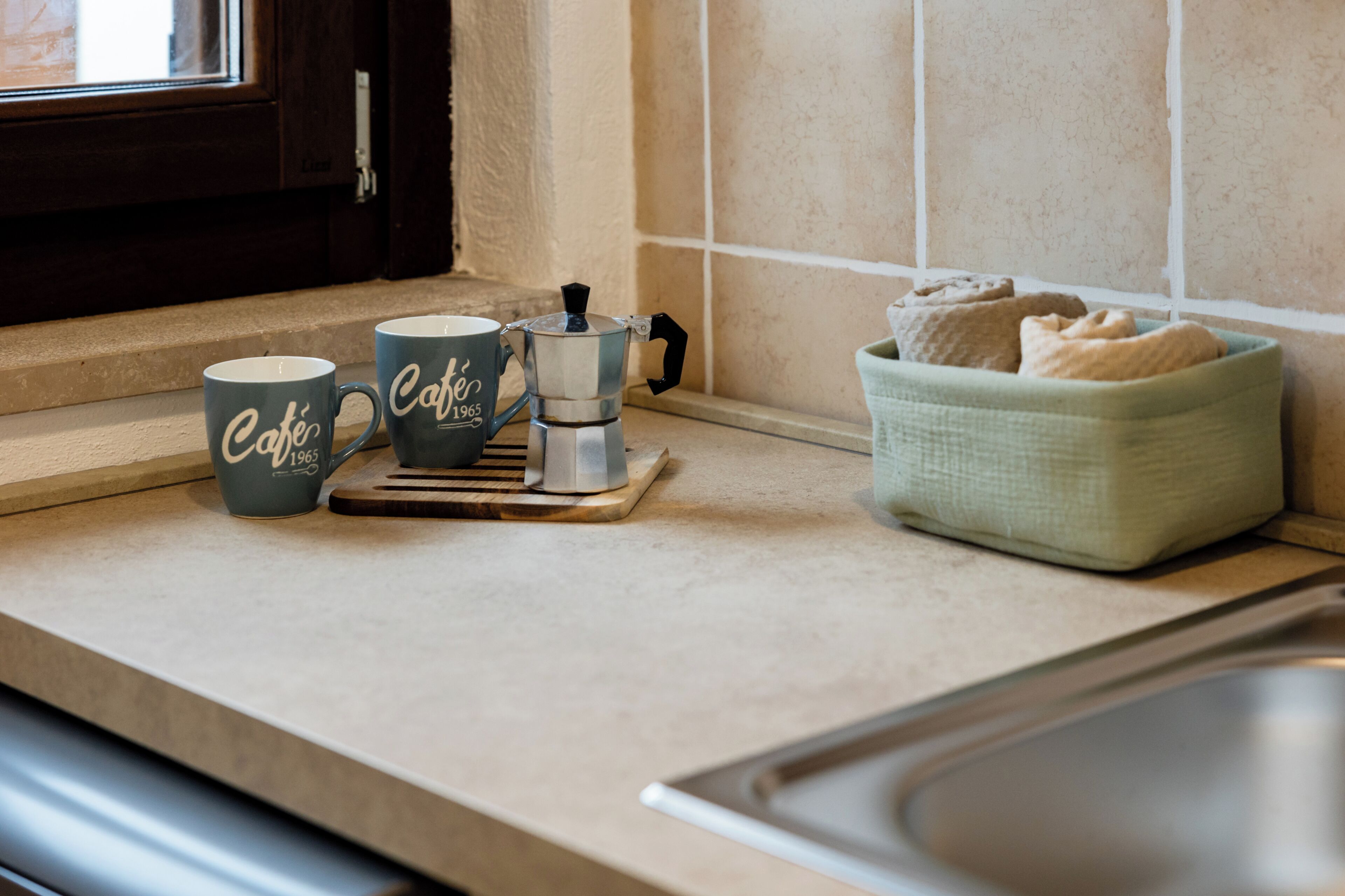A kitchen counter with two coffee mugs, a coffee pot, and a basket of bread.