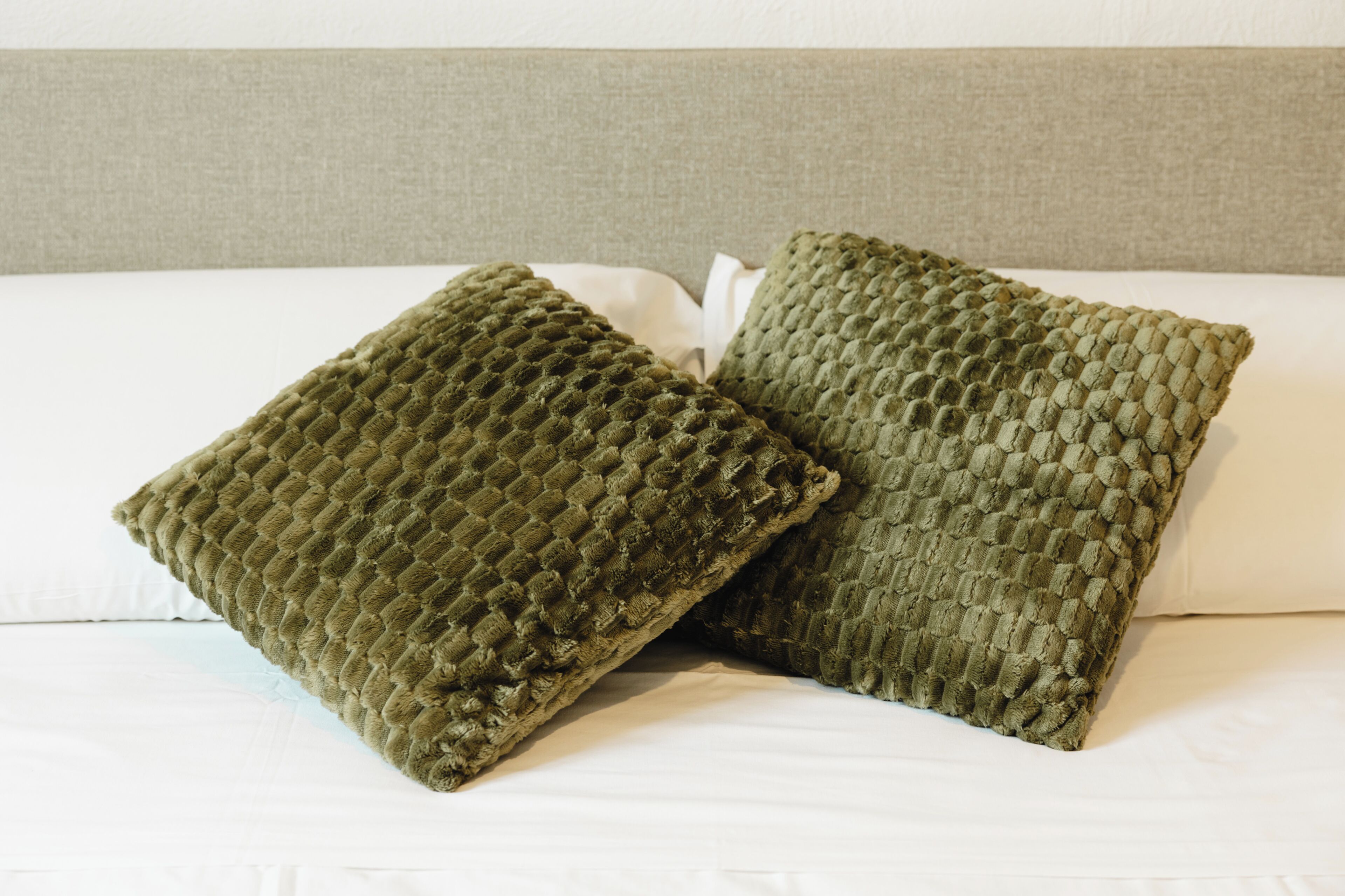 Deux coussins carrés à motifs texturés, posés sur un lit recouvert de draps blancs et doté d’une tête de lit grise.