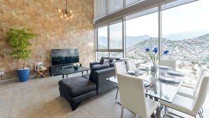 A modern living room with a large TV, comfortable seating, a dining area, and a city view through large windows. - Micrópolis (Monterrey)