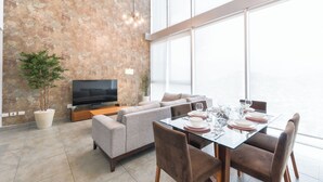 A modern living room with a flat-screen TV, a sofa, a dining table set for four, and a large window. - Micrópolis (Monterrey)