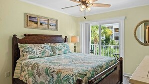 A bedroom with a wooden bed, a ceiling fan, a window with a view of greenery, and a framed picture on the wall. - Pelican's Landing 118- 3BR/2.5BA- Outdoor Pool (Myrtle Beach)