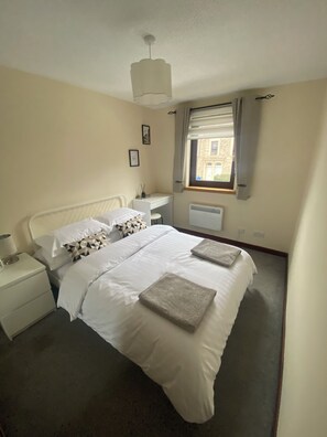 Une chambre à coucher comprenant un lit, des tables de chevet, une fenêtre avec des rideaux, un radiateur et un luminaire au plafond.