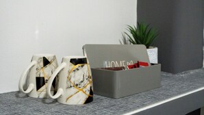 Dos tazas con diseño de mármol sobre una estantería, una caja gris con la palabra 'HOME' escrita y una pequeña planta en maceta al fondo.