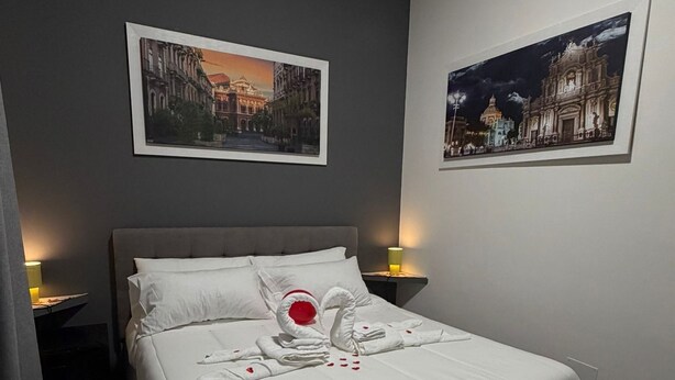 Una cama bien hecha con ropa de cama blanca y una toalla dispuesta en forma de cisne, dos cuadros en la pared y una lámpara de noche.