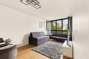 A modern living room with a grey sofa, a black sofa, a flat-screen TV, and a large window offering a view of buildings and greenery. - Bright & Comfortable 2BR near Le Marais - Paris 11 (Paris)