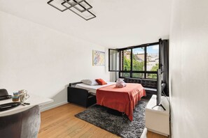 A compact room with a bed, a desk, a window with curtains, and a view of buildings outside. - Bright & Comfortable 2BR near Le Marais - Paris 11 (Paris)